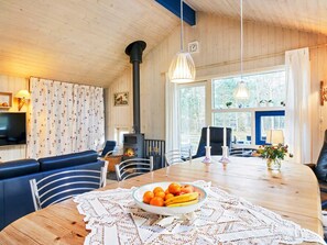 Interior acogedor de una cabaña con una mesa de comedor de madera, un cuenco de fruta, una chimenea y un ventanal grande.