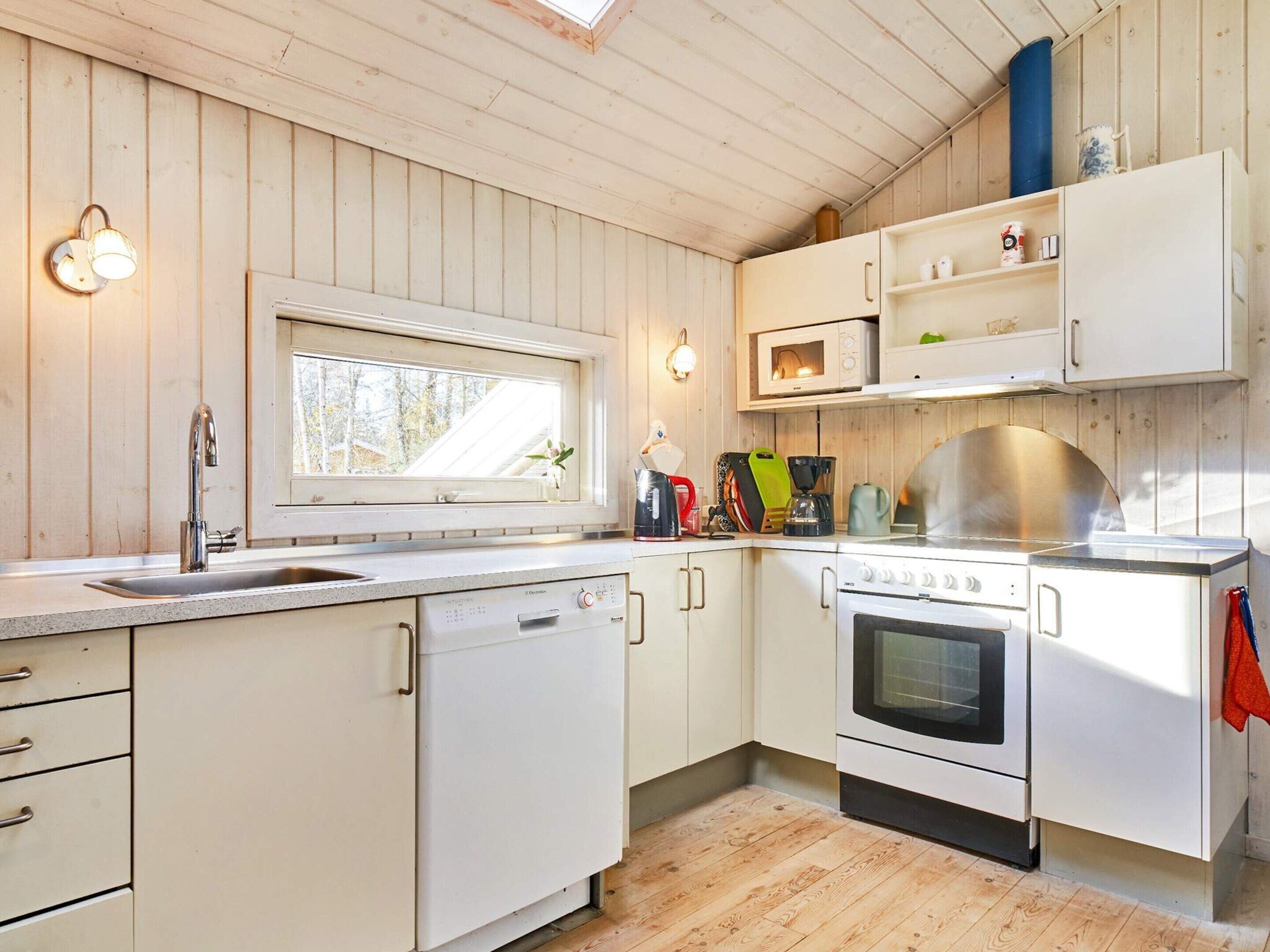 A kitchen with wooden panel walls, a window, a sink, a dishwasher, a microwave, a stove, and a shelf with kitchen items.