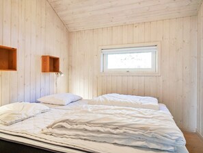 Un dormitorio con techo de listones de madera, una cama individual con ropa blanca, una ventana pequeña y un estante de madera.