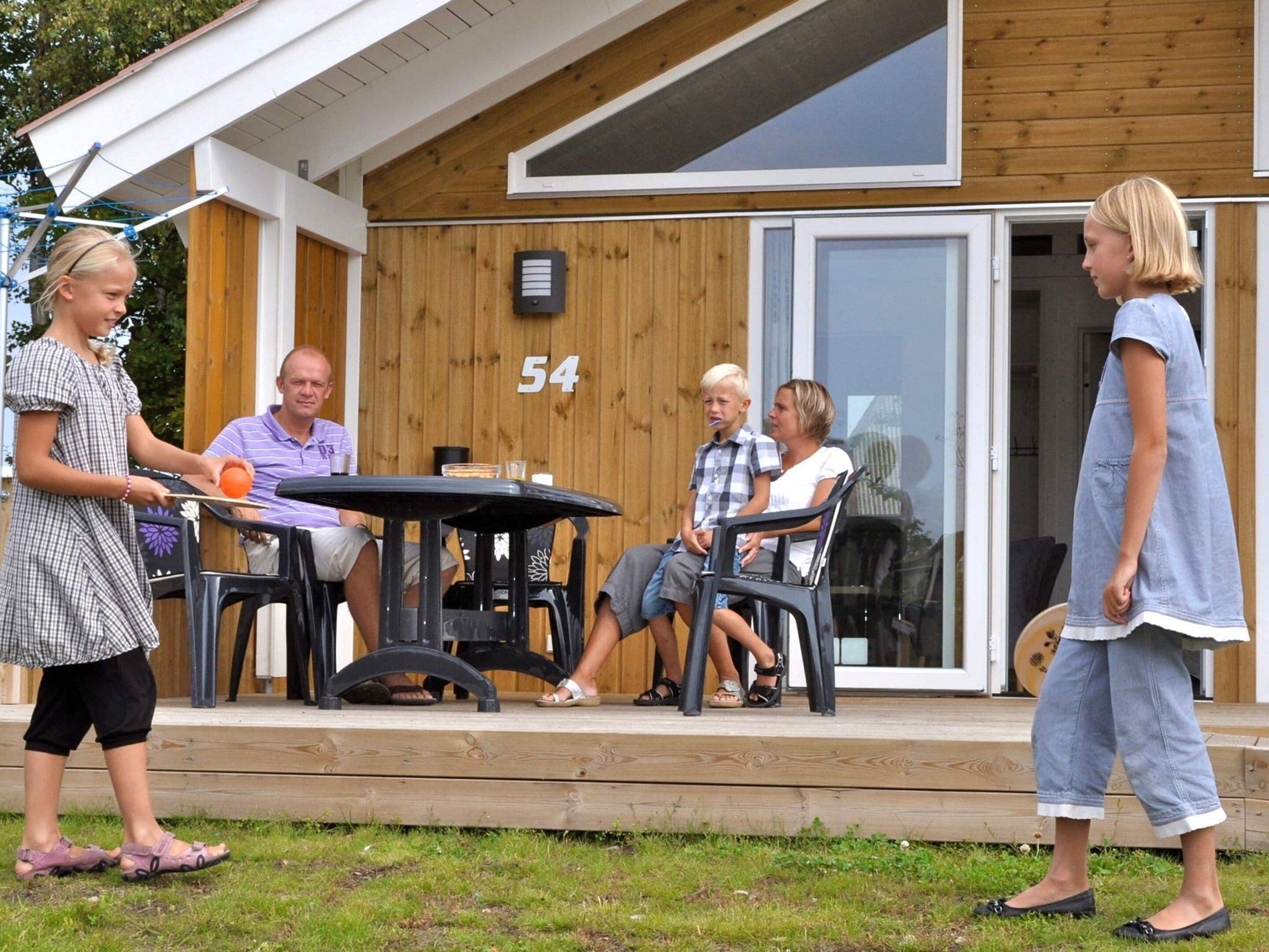 Ett trähus med siffran ’54’ på väggen, en uteplats med ett bord och stolar, och ett barn som leker med en boll.