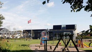 Un parc infantil amb cadira d'oscil·lació i estructura per escalar, envoltat d'una zona verda i un edifici amb una bandera.