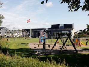 A playground with swings and a slide, a picnic table, and a building with a flag in the background. - 4 Star Holiday Home in Skaerbaek (Skaerbaek)
