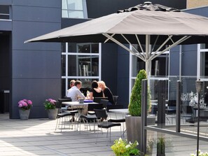 Een terras met tafels en stoelen onder een grote parasol, omgeven door potplanten en een modern gebouw op de achtergrond.