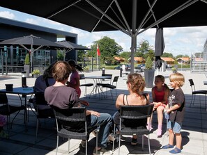 An outdoor seating area with tables and chairs under a shaded umbrella, a building in the background, and a clear sky. - 4 Star Holiday Home in Skaerbaek (Skaerbaek)