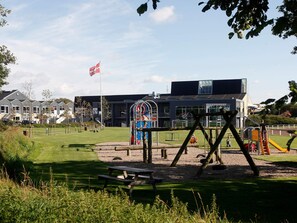 A playground with swings and a slide, a picnic table, and a building with a flag in the background. - 4 Star Holiday Home in Skaerbaek (Skaerbaek)
