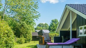 A wooden deck with a lounge chair and a striped umbrella, surrounded by greenery and a building with a sloped roof. - 4 Star Holiday Home in Borkop (Borkop)