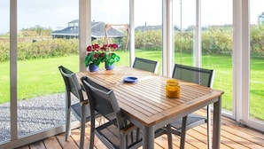 A covered patio with a wooden table and chairs, potted plants, and a view of a grassy yard and houses. - 5 Person Holiday Home in Lemvig-by Traum (Lemvig)