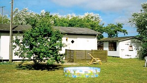 Una casa petita i blanca amb una tanca de fusta, un arbre i una piscina en un jardí amb gespa.