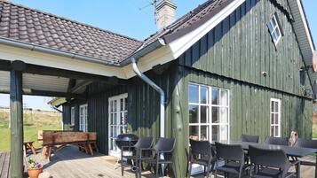 Una terraza de madera con mobiliario exterior y una parrilla, adosada a un edificio verde con tejado de tejas.