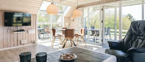 A modern living room with a flat-screen TV, a dining area with chairs, and a large window offering a view of the outdoors.