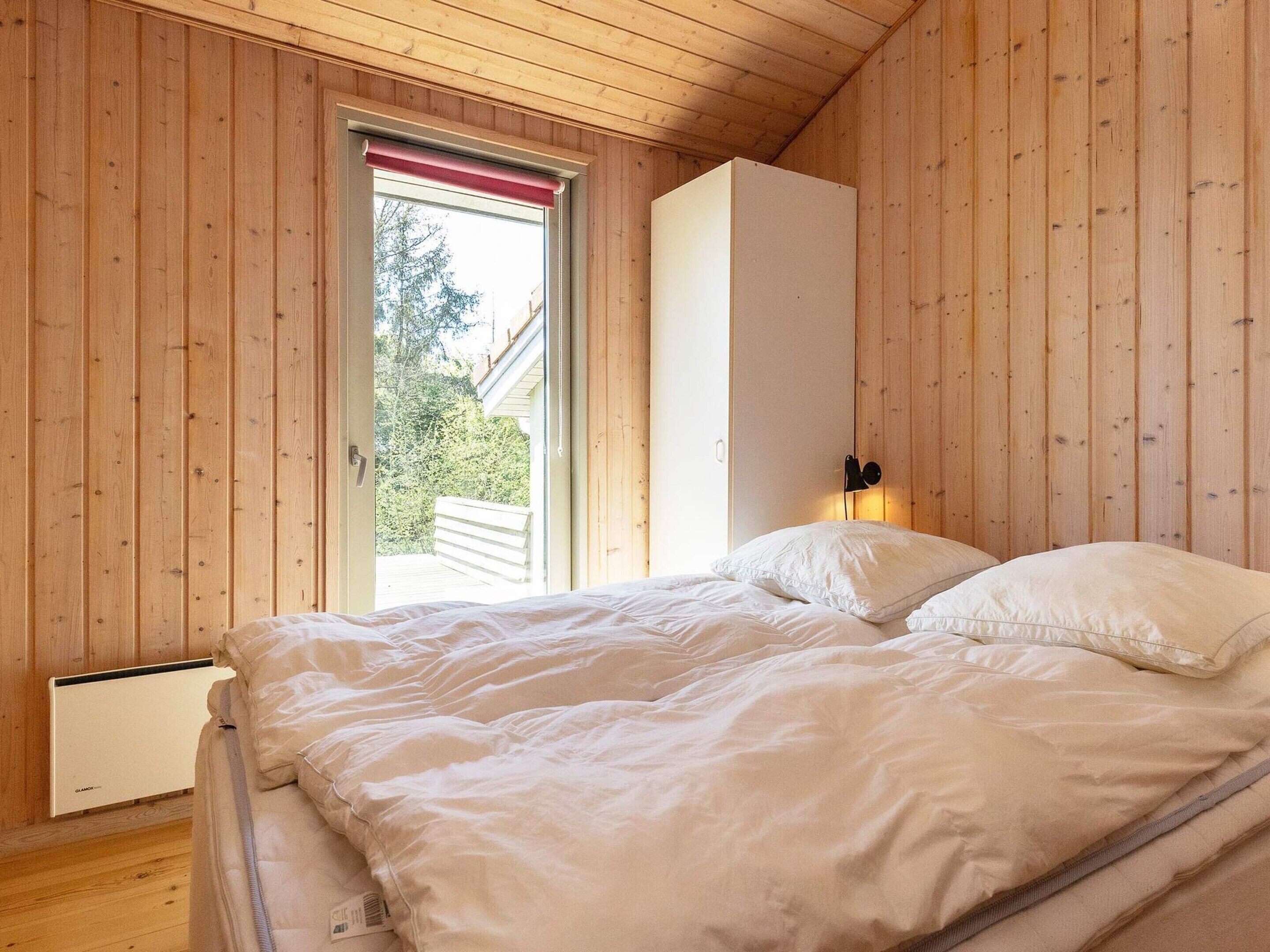 A bedroom with a wooden ceiling, a bed with white bedding, a radiator, and a window with a red blind.