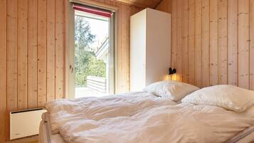 A bedroom with a wooden ceiling, a bed with white bedding, a radiator, and a window with a red blind.