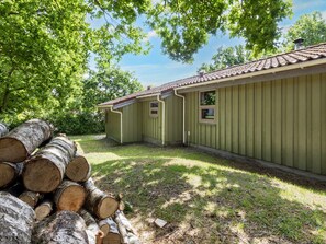 A row of green cabins with a tiled roof, surrounded by trees and a pile of logs.