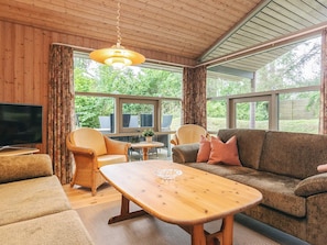 Un salon chaleureux avec un plafond en bois, une télévision, un canapé, une table basse et de grandes fenêtres donnant sur un paysage verdoyant.