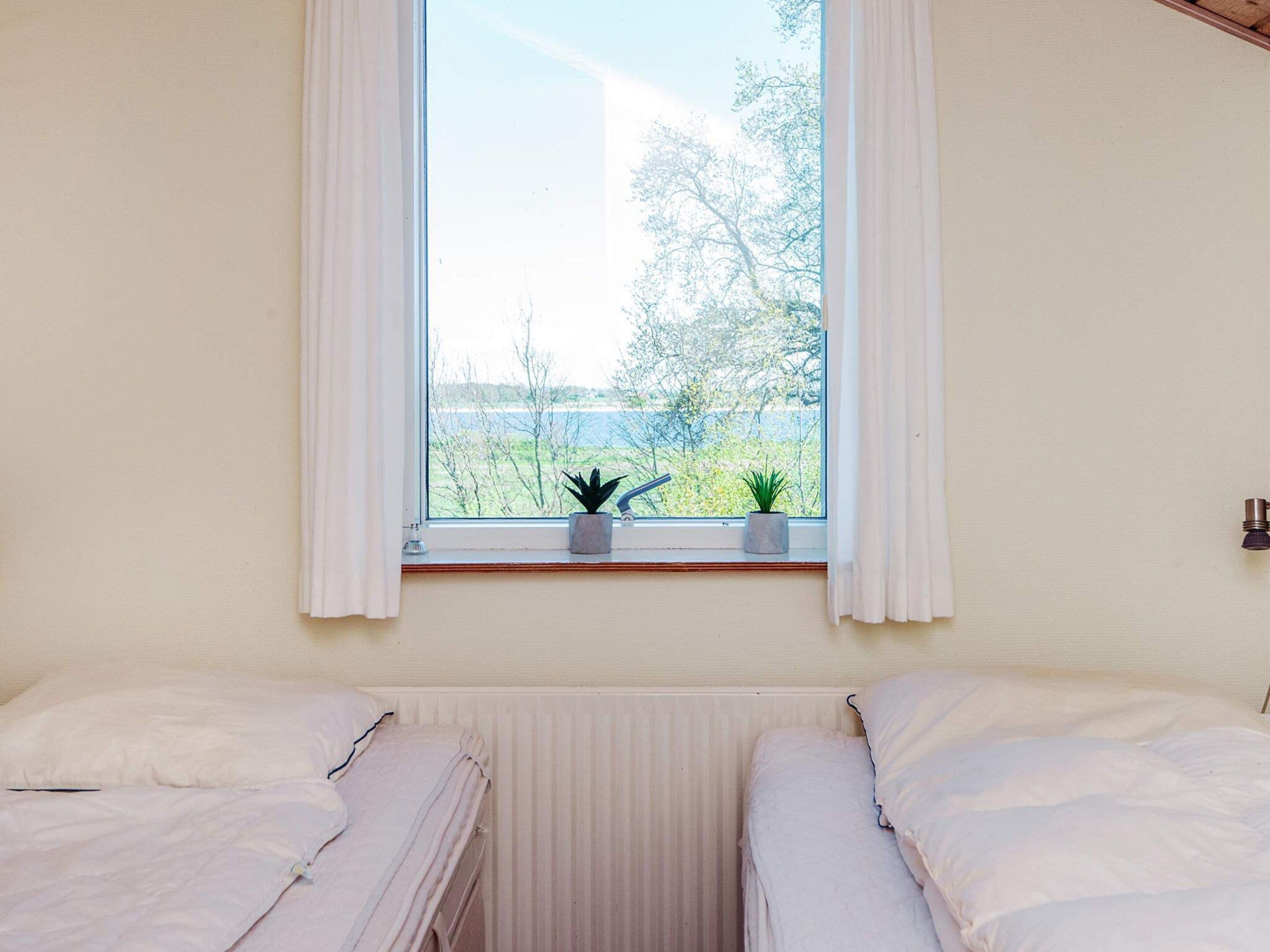 A room with two beds, a window with curtains, and two potted plants on the windowsill.