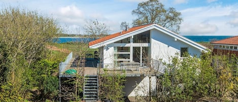 A modern house with a deck, surrounded by greenery and a view of the sea.