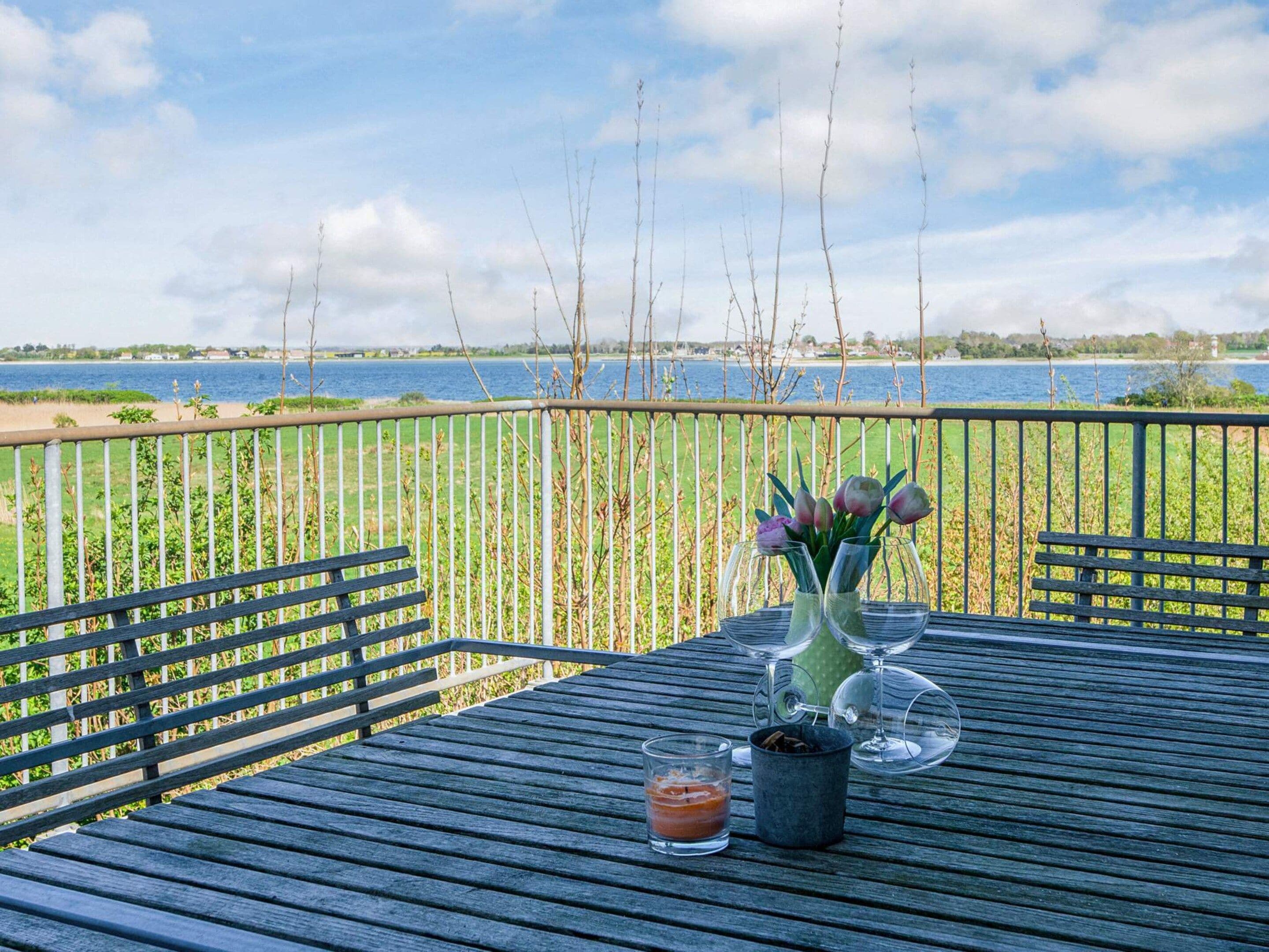 A wooden deck with a table set for two, overlooking a body of water and a grassy field.