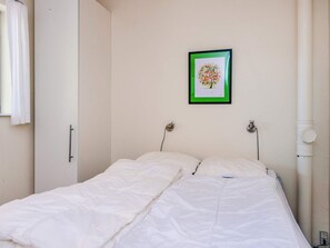 Una cama individual con ropa de cama blanca, un cuadro colgado en la pared con motivos florales, y una tubería blanca en el lado derecho.