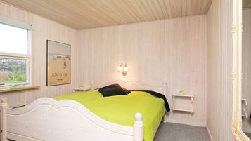 A bedroom with a white bed, a green bedspread, a framed picture on the wall, and a bedside table with a lamp.
