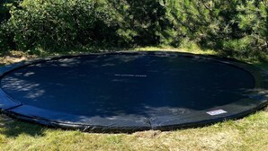 Ein runder Trampolin in einer grünen Wiese, im Hintergrund dichte Bäume.