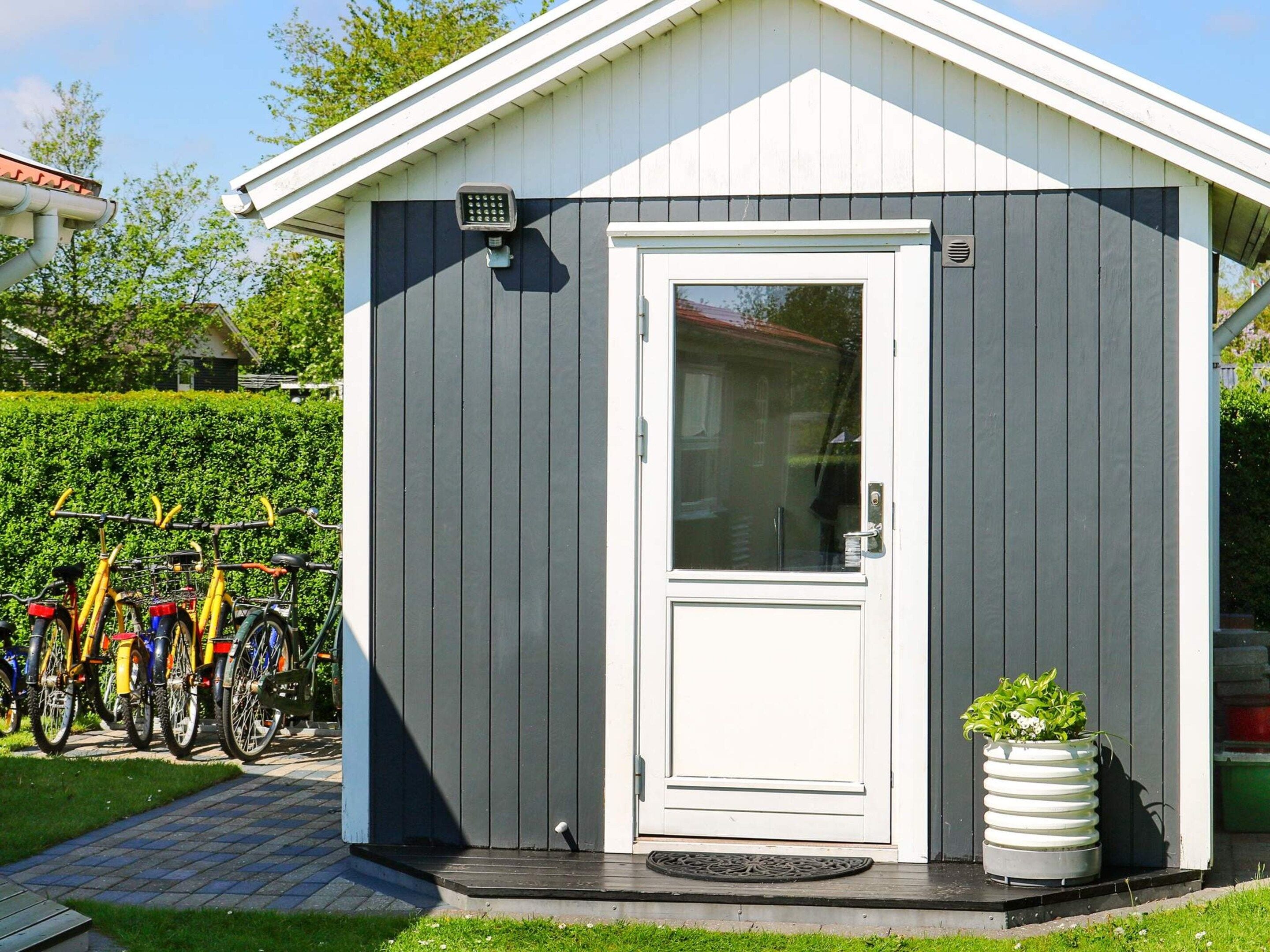 Een klein tuinhuisje met een witte deur en een raam, een fiets geparkeerd ervoor en een bloempot naast de deur.
