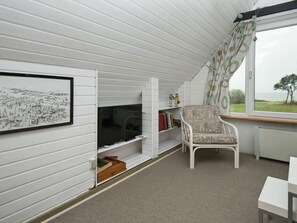 A room with a white interior, a fireplace, a television, a chair, a table, and a window with a view of the sea. - Cozy Holiday Home in Hemmet near Sea (Hemmet)