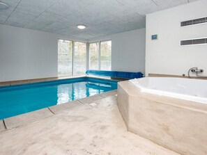 Une piscine intérieure avec un espace jacuzzi séparé, de grandes fenêtres et un sol carrelé.