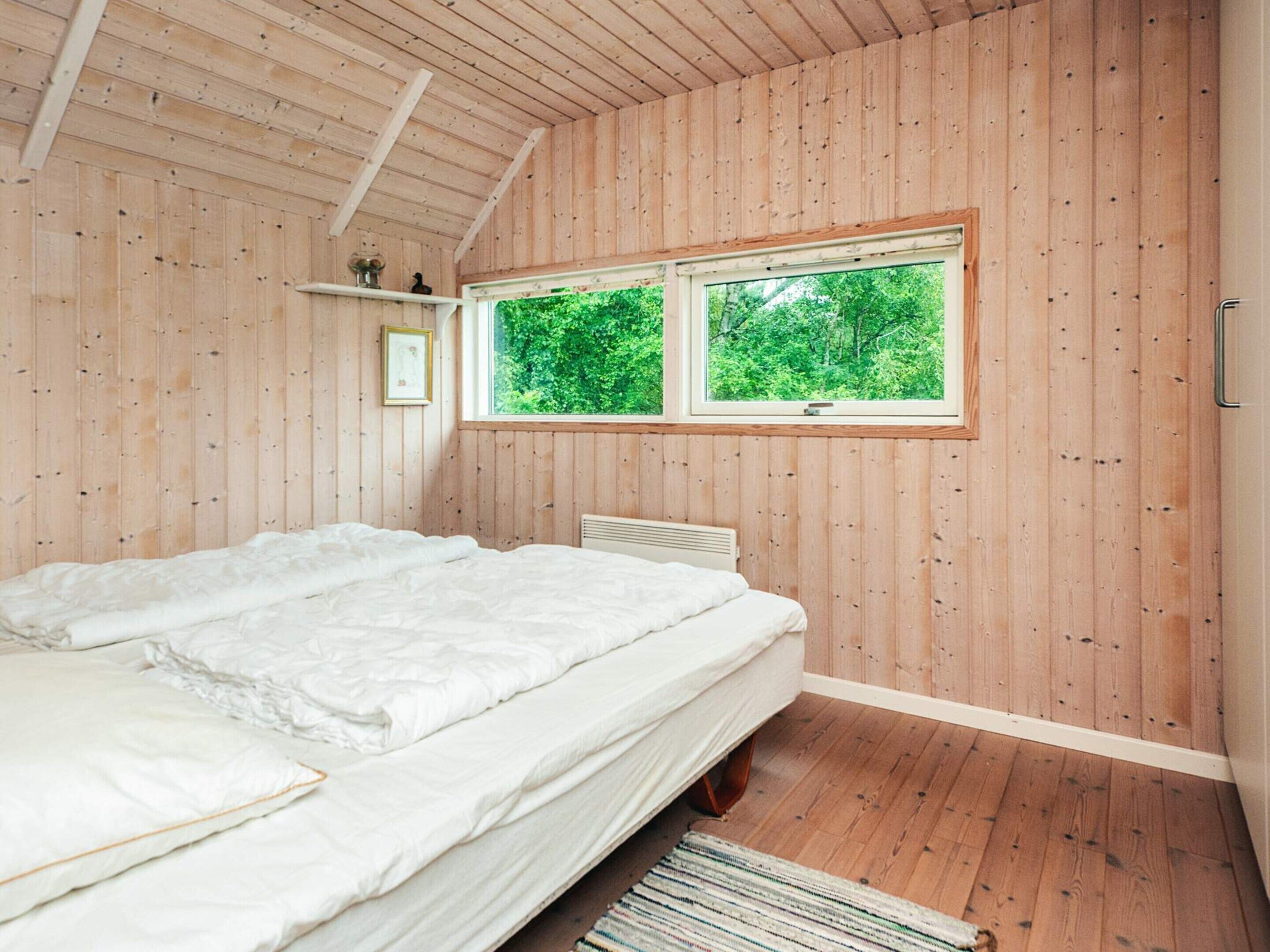 A bedroom with wooden walls, a large window, a bed with white bedding, and a small shelf with decorative items.