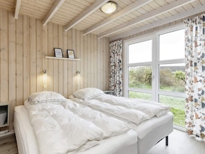 Quarto com duas camas, paredes de madeira, janela com cortinas e prateleira com quadros emoldurados.