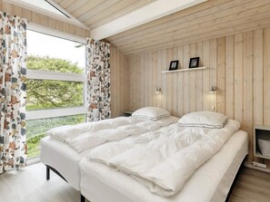A bedroom with a wooden ceiling, a bed with white bedding, a window with floral curtains, and a small shelf with picture frames. - 5 Star Holiday Home in Hjorring (Hjørring)