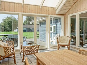A covered patio area with a wooden table and wicker chairs, large glass doors leading to a backyard, and a view of a house in the distance. - 4 Star Holiday Home in Aakirkeby (Akirkeby)