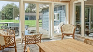 Een overdekt terras met een houten tafel en rieten stoelen, grote glazen deuren die uitkomen op een achtertuin, en een uitzicht op een huis op de achtergrond.