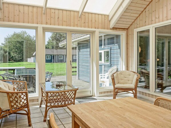 A covered patio with a wooden table and wicker chairs, large glass doors leading to a backyard, and a view of a house in the background.