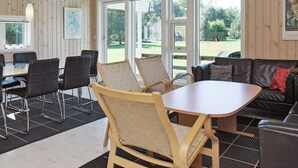 Een kamer met een houten inrichting, een ronde tafel en een bank. Er staan stoelen rondom de tafel en een raam met uitzicht op groen.