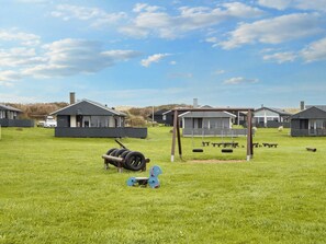 A grassy field with a swing set, tire swings, and a small blue chair. There are several houses with dark roofs in the background. - 5 Person Holiday Home in Lemvig (Lemvig)