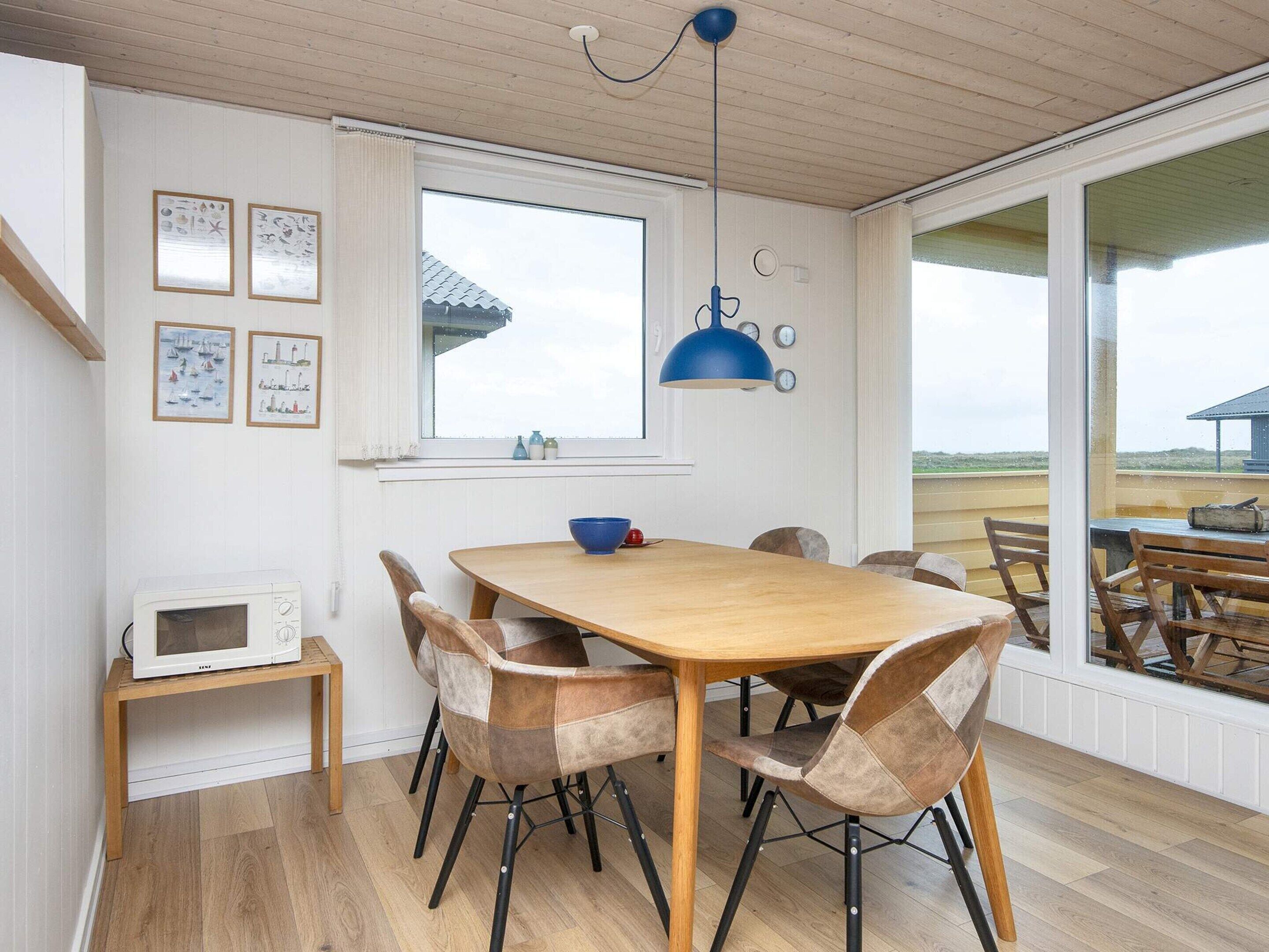 Un espace repas avec une table et des chaises en bois, un luminaire suspendu bleu et une vue sur l’extérieur.
