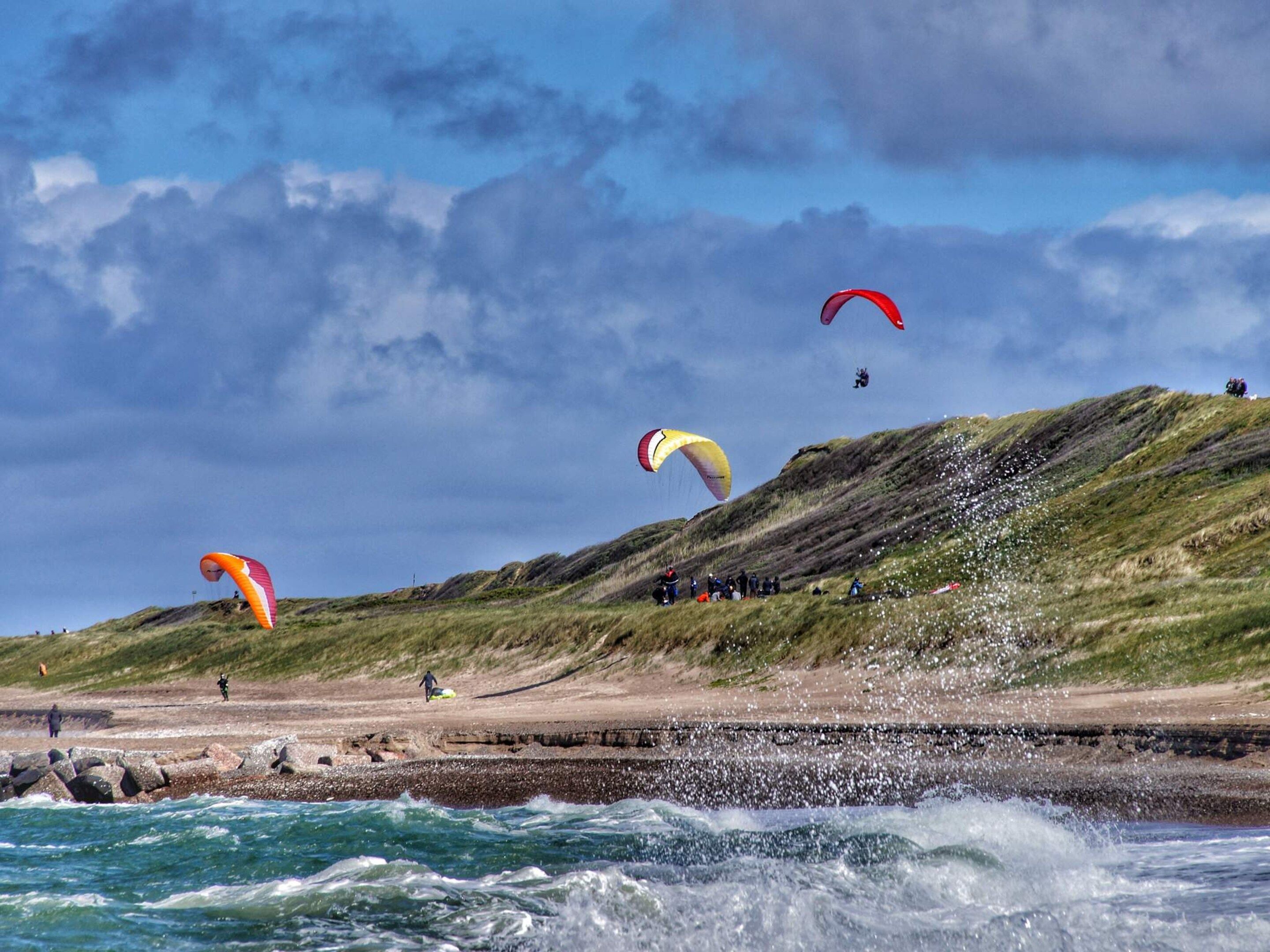 A coastal scene with people engaging in kite surfing, a rocky shoreline, and waves crashing onto the beach.