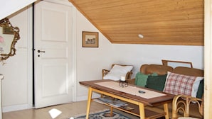 A cozy room with a wooden ceiling, a white door, a framed picture on the wall, a wooden table with a cushion, and a patterned rug.