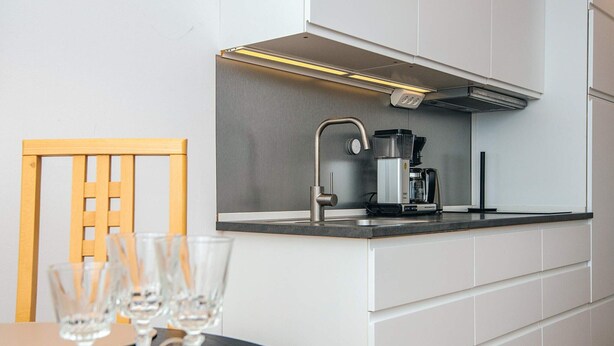 A modern kitchen with a built-in stove, a coffee maker, and a sink. There is a wooden chair and a table with glasses on it. - 4 Person Holiday Home in Romo (Rømø)