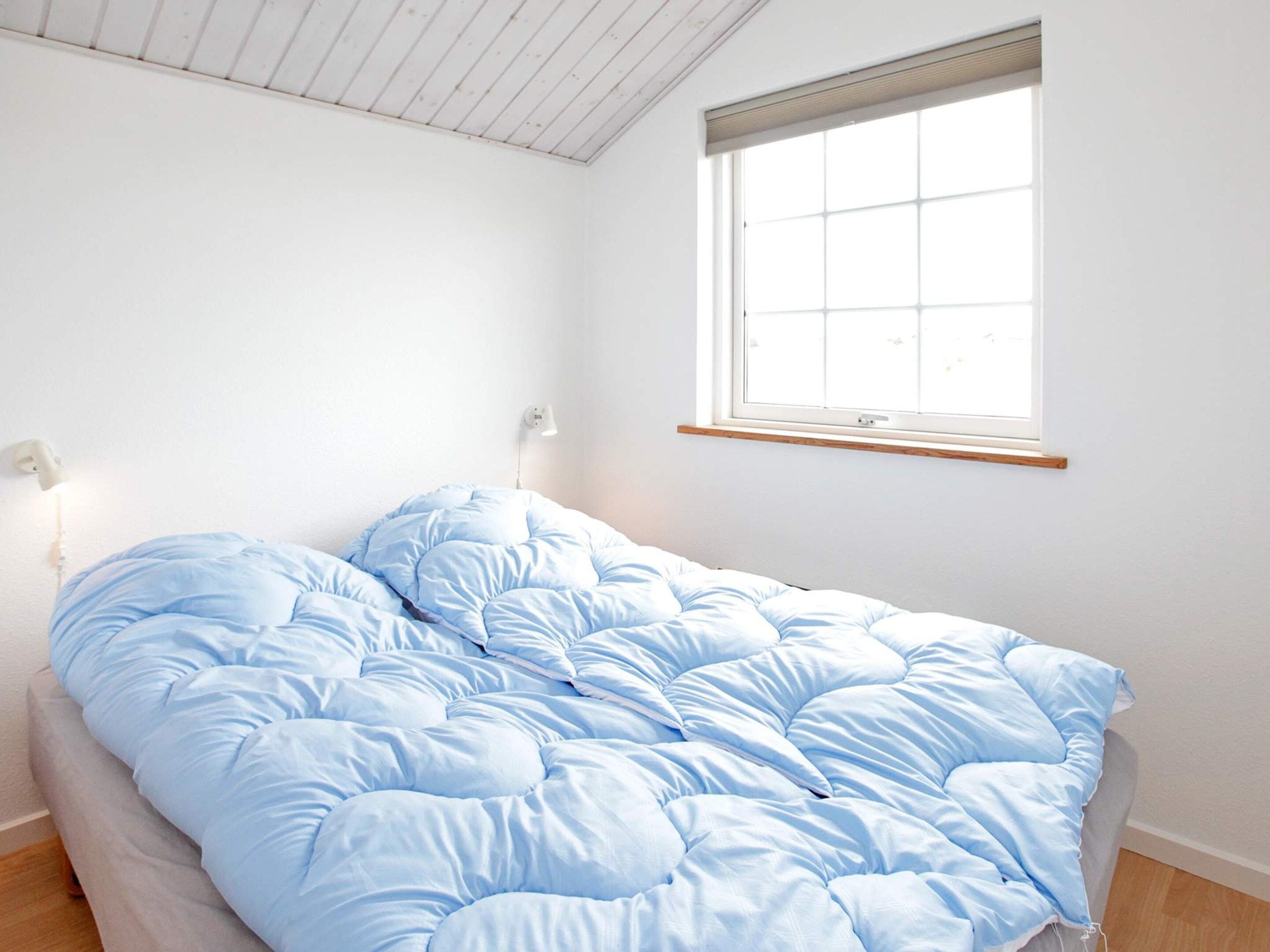 A single bed with a blue duvet in a room with white walls, a window, and a wall-mounted light fixture.