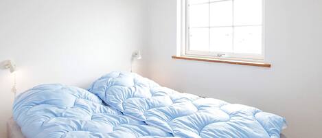A single bed with a blue duvet in a room with white walls, a window, and a wall-mounted light fixture.