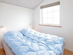 A single bed with a blue duvet in a room with white walls, a window, and a wall-mounted light fixture.