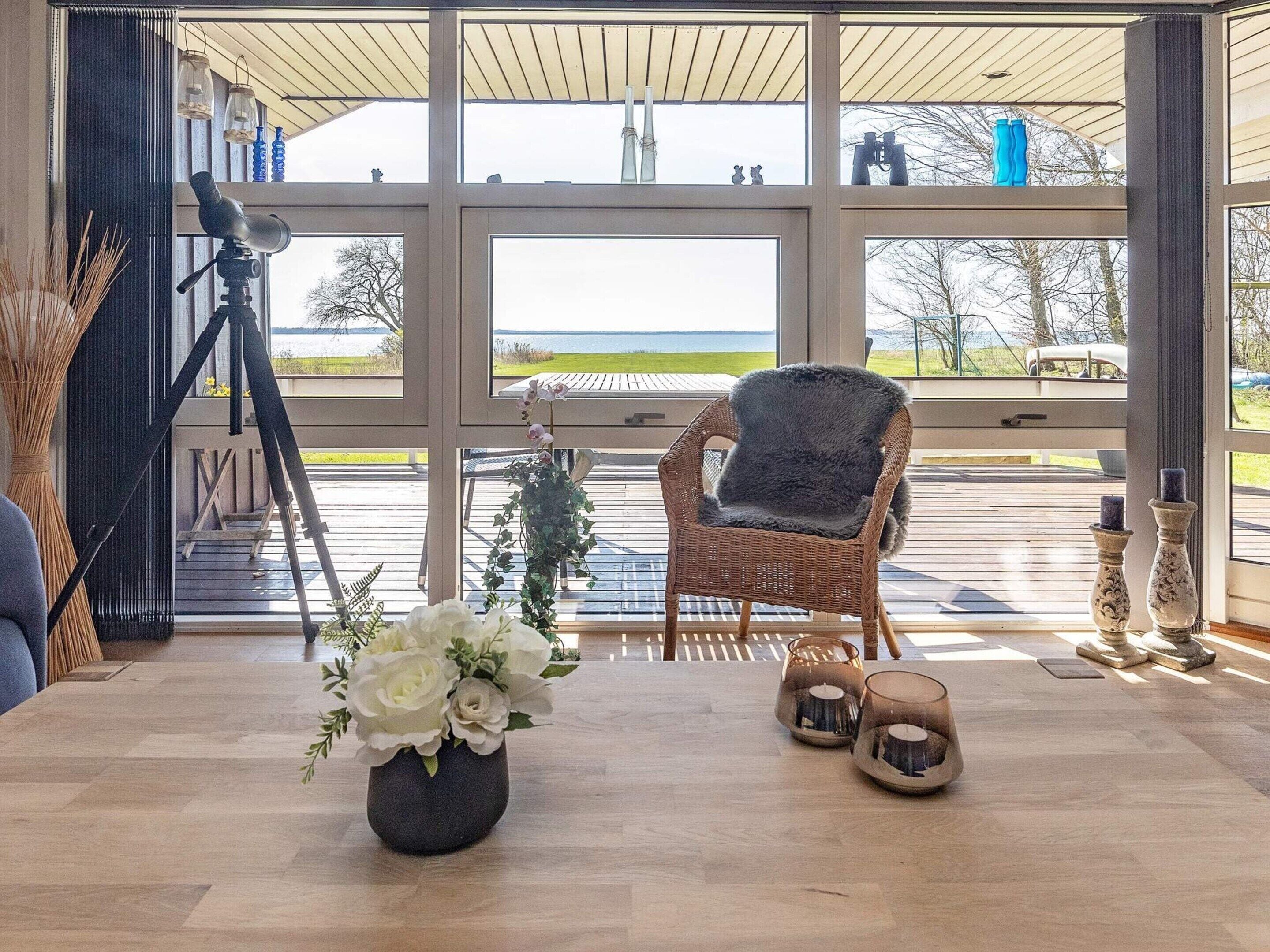 Een kamer met een groot raam, een rieten stoel, een statief en een vaas met bloemen.