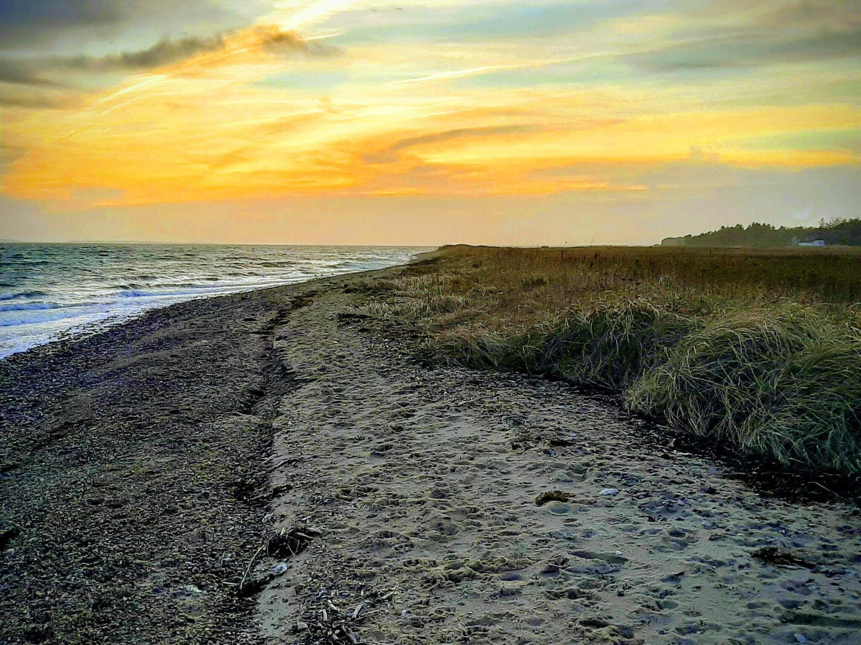Uma praia com um caminho de areia que leva à água, dunas gramadas e um pôr do sol ao fundo.