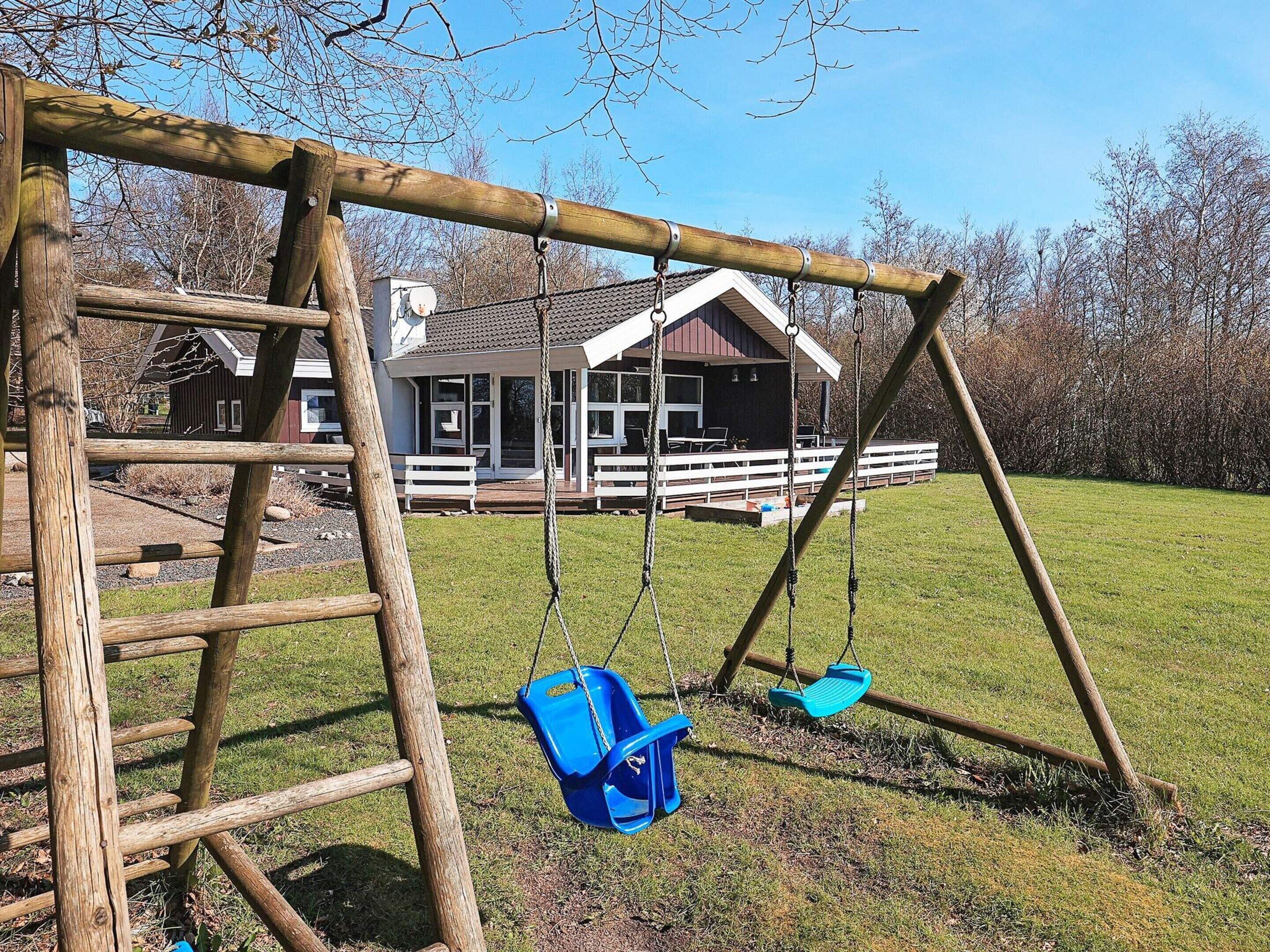 Une balançoire en bois avec deux sièges et une échelle, placée devant un décor représentant une maison et un champ d’herbe.