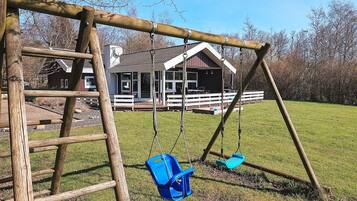 Une balançoire en bois avec deux sièges et une échelle, placée devant un décor représentant une maison et un champ d’herbe.