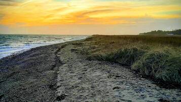 Una playa con un sendero arenoso que lleva al agua, dunas cubiertas de hierba y una puesta de sol al fondo.