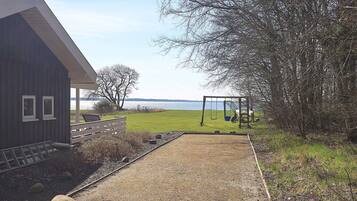 A house with a gravel path leading to a swing set, a fenced area, and a view of a lake.