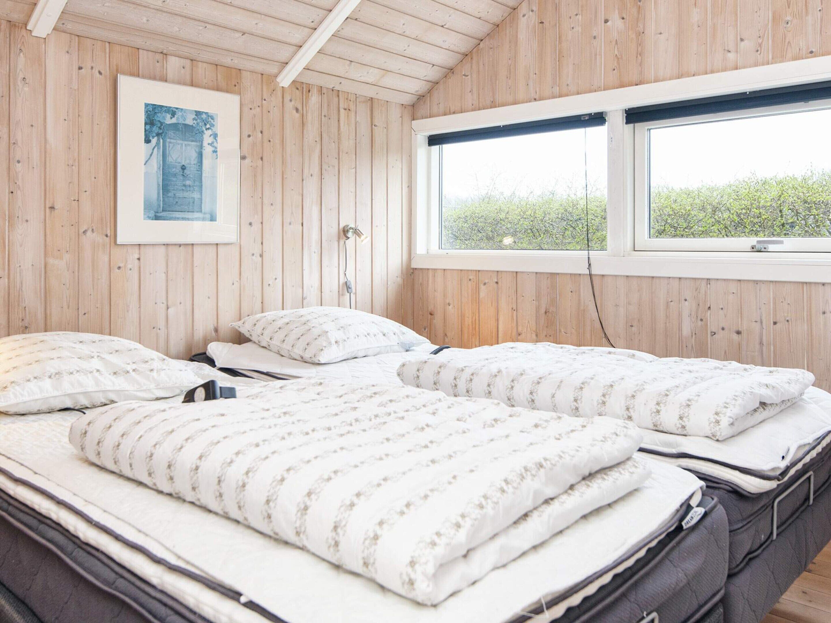A room with wooden walls, a bed with a white floral bedspread, a window with a view of greenery, and a framed picture on the wall.