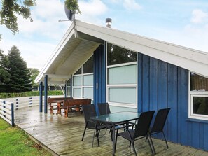 A covered outdoor area with a table and chairs, a wooden deck, and a blue building with large windows.